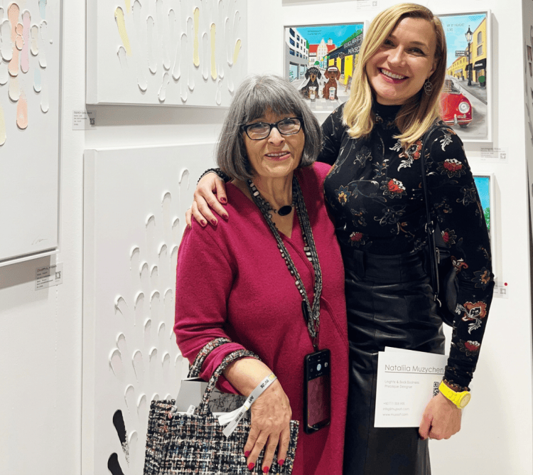 Zwei Frauen posieren in einem Kunstraum vor weißen, reliefartigen Kunstwerken. Die ältere Frau trägt ein lila Oberteil und eine Handtasche, während die jüngere Frau schwarze Kleidung mit floralem Muster trägt und lächelt.