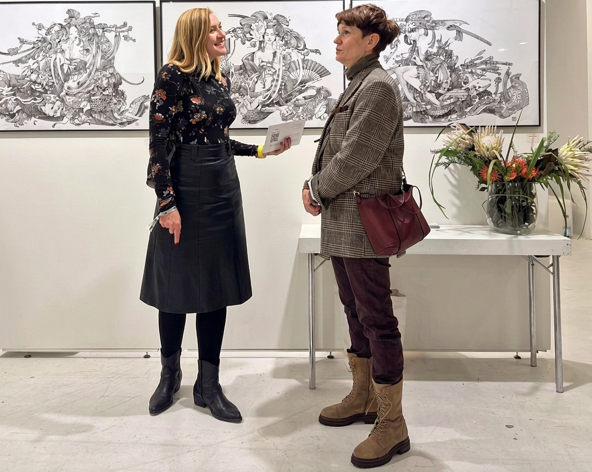WhatsApp Image 2026-10-23 at 20.07.39 (1) Zwei Frauen stehen in einem Kunstraum, während sie miteinander sprechen. Im Hintergrund sind großformatige Zeichnungen zu sehen. Eine Frau trägt eine schwarze Bluse mit floralen Motiven und einen schwarzen Lederrock, während die andere Frau einen karierten Blazer und braune Stiefel trägt. Ein Blumengefäß steht auf einem Tisch zwischen ihnen.