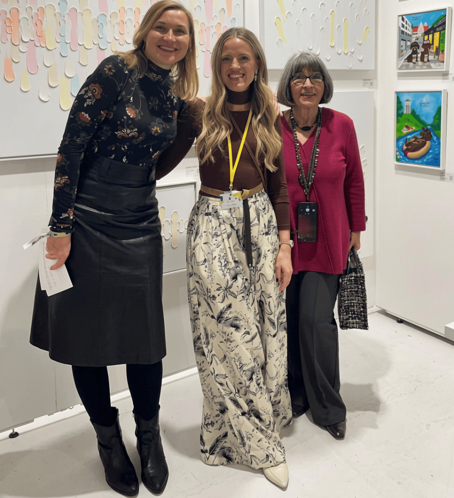 Drei Frauen posieren vor einer Kunstinstallation in einem Ausstellungsraum. Eine Frau trägt ein schwarzes Oberteil und einen langen Rock, eine andere trägt ein braunes Oberteil mit einer langen, bedruckten Hose, und die dritte trägt ein rotes Oberteil und hat eine Tasche. Im Hintergrund sind farbige Kunstwerke sichtbar.
