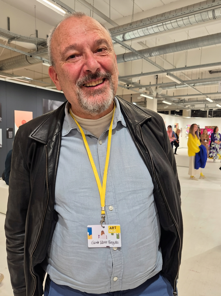 20261023_184603 Ein lächelnder Mann mit Bart und kahlem Kopf trägt eine Lederjacke und ein hellblaues Hemd. Er steht in einer modernen Galerie mit Kunstwerken im Hintergrund.
