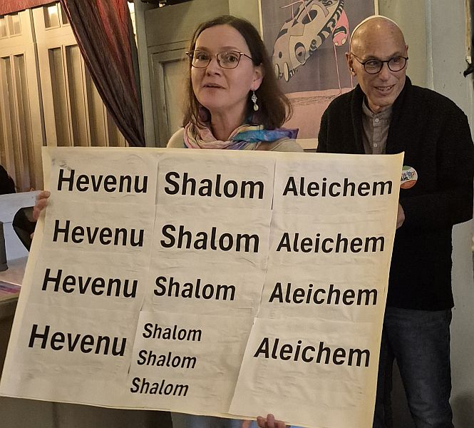 Eine Frau hält ein großes Schild mit der Aufschrift 'Hevenu Shalom Aleichem' in verschiedenen Schriftgrößen. Im Hintergrund steht ein Mann, der lächelt.