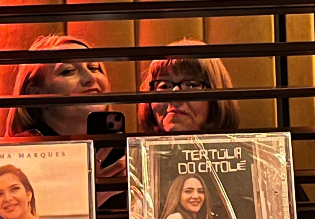 Zwei Frauen, die hinter einem Gitter stehen, lächeln in die Kamera. Im Hintergrund sind verschiedene Plakate zu sehen.