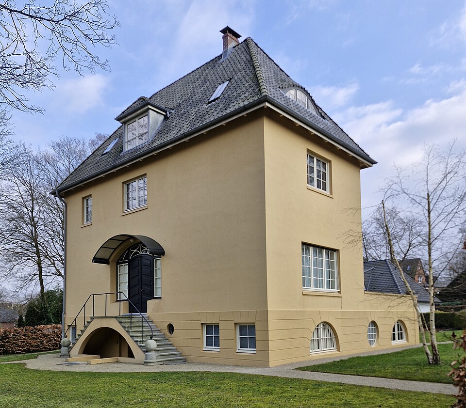 Ein modernes, dreigeschossiges Haus mit einer gelben Fassade, einer gewölbten Eingangstreppe und einem Satteldach. Umgeben von einem gepflegten Garten und vereinzelten Bäumen.